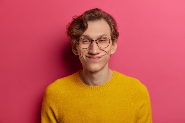 Headshot van grappige positieve hipster-man lacht vrolijk, heeft oprechte optimistische blik, draagt een ronde transparante bril en een gele trui, luistert naar een hilarisch verhaal, geïsoleerd op een roze muur