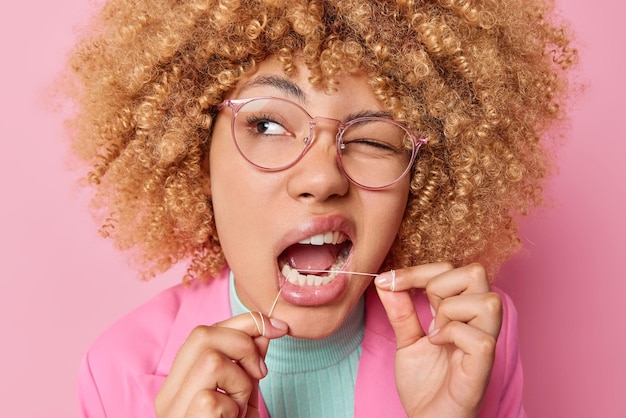 Gratis foto headshot van jonge vrouw met krullend haar gebruikt tandzijde knipoogt oog en zorgt voor mondhygiëne geconcentreerd opzij draagt transparante bril elegante kleding geïsoleerd over roze achtergrond