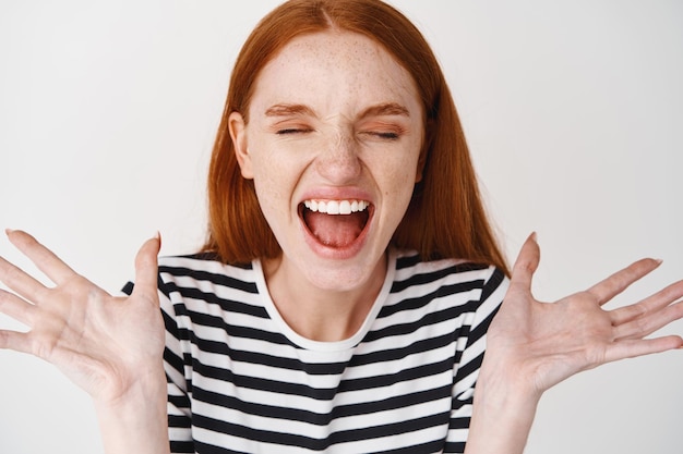 Headshot van jonge vrouw met rood haar, sproeten en bleke huid schreeuw van vreugde, zich gelukkig en extatisch voelen, handen zijwaarts spreiden en juichen van vreugde