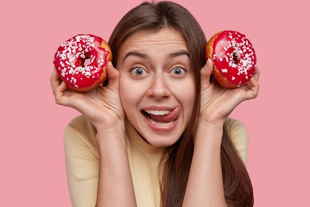 Headshot van knappe jonge dame likt lippen, kijkt met vreugdevolle uitdrukking, draagt twee smakelijke donuts, heeft donker haar, geniet van snack