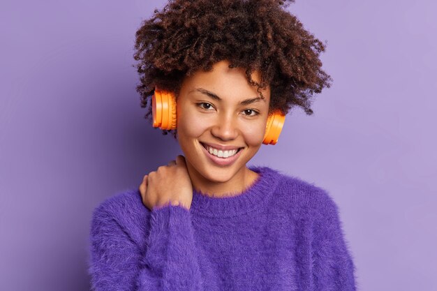 Headshot van knappe krullende harige vrouw met Afro haar glimlach zachtjes raakt nek kijkt positief naar camera luistert audiotrack via draadloze stereo hoofdtelefoon