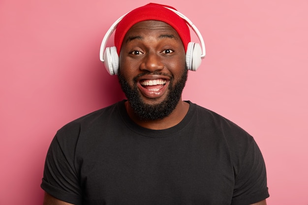 Headshot van knappe man met brede glimlach, heeft witte tanden, levert geluid aan oren via koptelefoon, luistert naar muziek in draadloze hoofdtelefoon