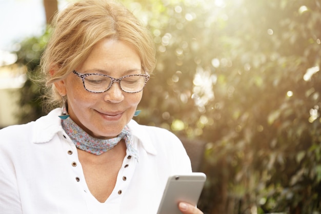 Headshot van knappe moderne blonde volwassen vrouw die een bril draagt, haar kleinzoon een bericht stuurt via sociale netwerken, haar generieke mobiele telefoon gebruikt, lacht terwijl ze een bericht leest of naar de foto kijkt