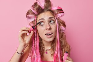 Headshot van langharige jonge europese vrouw past haarrollers toe en mascara kijkt weg ondergaat schoonheidsbehandelingen bereidt zich voor op eerste date wil er mooi uitzien geïsoleerd over roze achtergrond