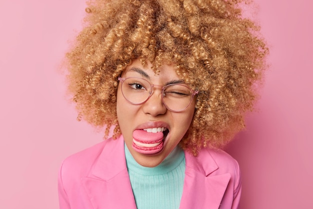 Headshot van mooie vrouw met krullend borstelig haar houdt heerlijke lekkere donut in mond knipoogt oog draagt transparante bril en formele jas geïsoleerd over roze achtergrond Suikerdieet of verslaving