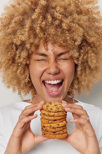 Gratis foto headshot van optimistische vrouw houdt stapel heerlijke koekjes met chocolade roept uit en voelt zich erg gelukkig houdt ogen gesloten mond wijd geopend, nonchalant gekleed lekker dessert en voeding
