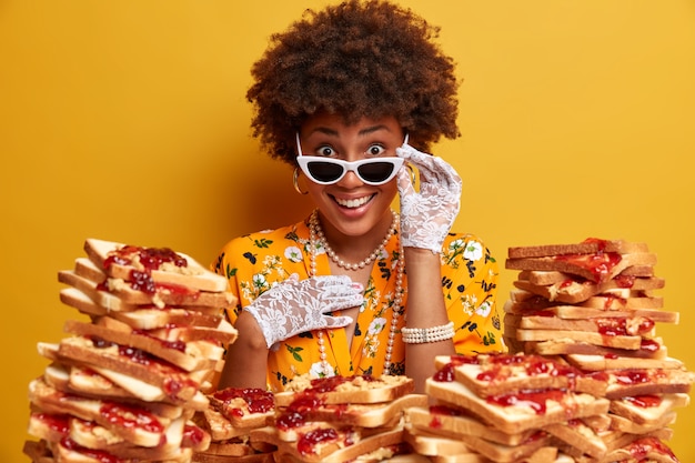 Headshot van positieve gekrulde afro-amerikaanse vrouw kijkt onder zonnebril, gekleed in modieuze kleding met ketting, besteedt vrije tijd aan het bezoeken van café, eet heerlijke toast met jam