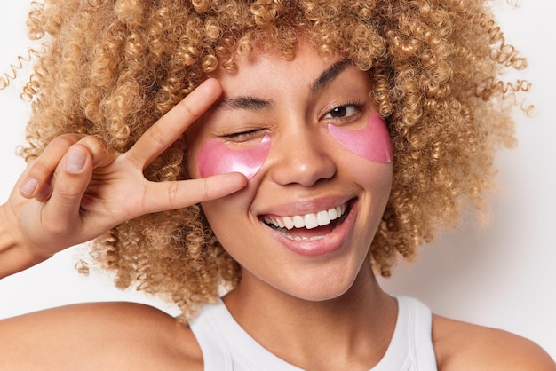 Headshot van positieve vrouw met krullend haar glimlacht breed knipoogt ogen toont vredesteken ondergaat schoonheidsprocedures past hydrogel pads toe onder ogen geïsoleerd op witte achtergrond. Huidverzorgingsconcept