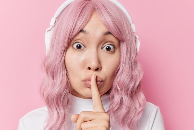 Headshot van verrast roze harige jonge Aziatische vrouw deelt geheim houdt wijsvinger over lippen vraagt om stil te zijn heeft wijd geopende ogen draagt draadloze stereo hoofdtelefoon luistert muziek poses binnen
