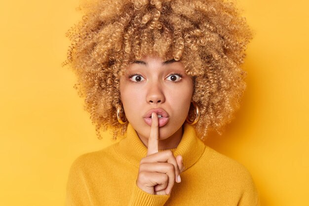 Headshot van verraste jonge vrouw met krullend haar drukt wijsvinger over lippen maakt stil gebaar kijkt mysterieus naar camera draagt trui geïsoleerd over levendige gele achtergrond vertelt geheim