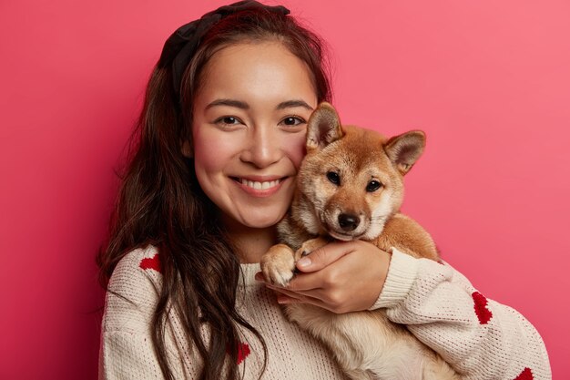 Headshot van vrolijke brunette jonge vrouw adopteert kleine pup, omhelst rashond, lacht aangenaam, geeft om huisdier, draagt warme trui