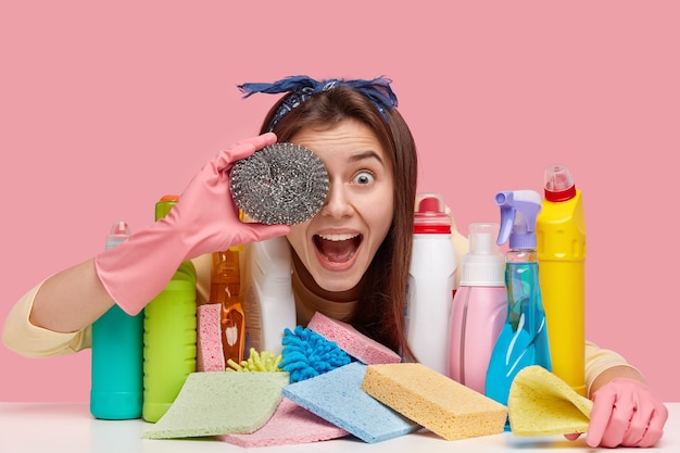 Headshot van vrolijke vrouw heeft donker steil haar heeft betrekking op oog met spons, heeft plezier na het schoonmaken van de kamer, vormt aan tafel met chemische producten in kleurrijke flessen