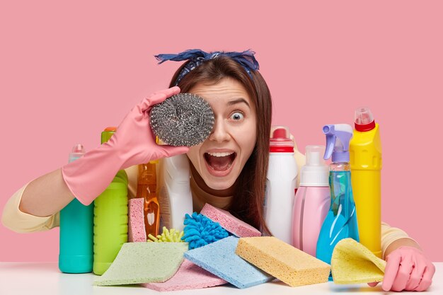 Headshot van vrolijke vrouw heeft donker steil haar heeft betrekking op oog met spons, heeft plezier na het schoonmaken van de kamer, vormt aan tafel met chemische producten in kleurrijke flessen