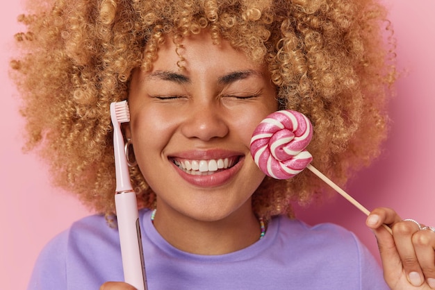 Headshot van vrolijke vrouw met krullend haar houdt ogen gesloten glimlacht breed heeft witte zelfs tanden houdt karamel snoep en elektrische tandenborstel poses tegen roze achtergrond schadelijk voedsel concept