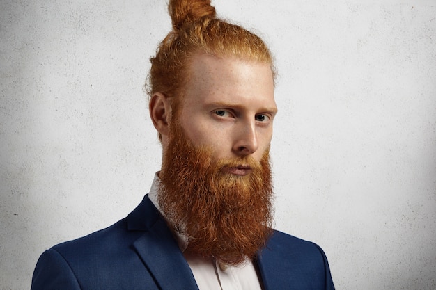 Gratis foto headshot van zelfverzekerde ondernemer met vage rode baard en haarknoop die zich in bureau op witte muur bevindt.