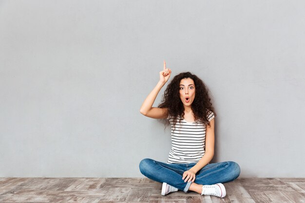 Heb een idee! Leuke vrouw die in vrijetijdskleding met benen zitten die op de vloer worden gesturing die wijsvinger omhoog betekenen die eureka over grijze muur betekenen