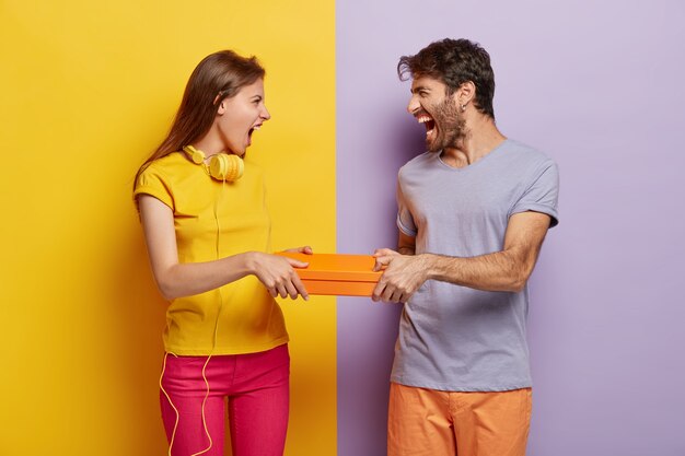 Hebzuchtige vrouw en man kunnen de doos niet delen, beide houden een oranje pakket vast, schreeuwen naar elkaar, hebben geïrriteerde uitdrukkingen, dragen levendige kleurrijke kleding, staan tegen een twee gekleurde achtergrond.