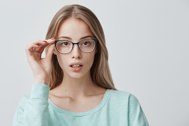 Heerlijke mooie blonde vrouw in trendy bril, draagt lichtblauwe trui, staat binnenshuis