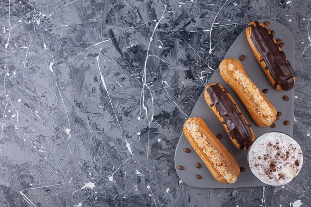 Heerlijke zoete chocolade en vanille eclairs en met een kopje koffie op een donkere snijplank.