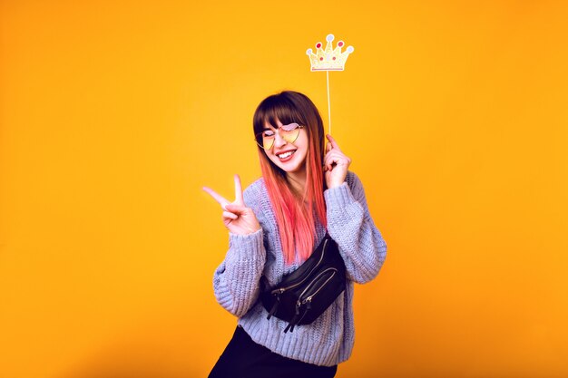 Helder grappig portret van vrolijke hipster vrouw met felroze haar, gezellige trui dragen, nep feestkroon vasthouden en glimlachen, klaar voor feest, gele muur.