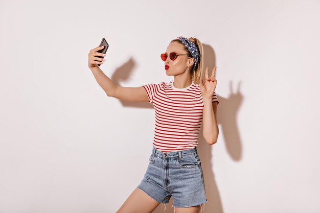 Helder meisje met rode lippen en zonnebril in gestreepte t-shirt en denim coole shorts die vredesteken tonen en selfie maken op witte achtergrond