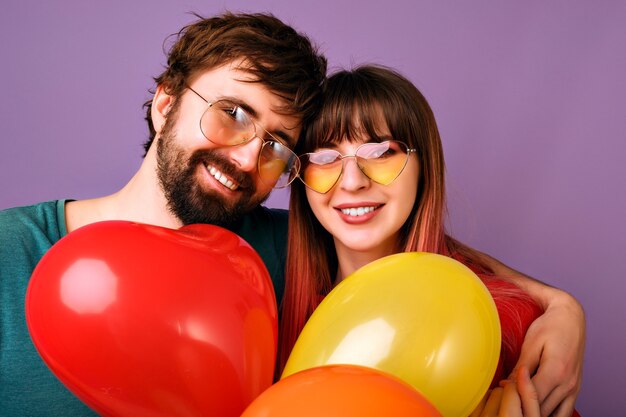 Helder positief portret van het gelukkige mooie paar glimlachen, vredesgebaar tonen en partijballons, familierelatie, violette muur houden