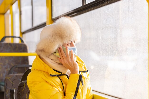 Helder zonnig portret van een jonge vrouw in warme kleren in een stadsbus op een winterse dag met een mobiele telefoon in haar hand