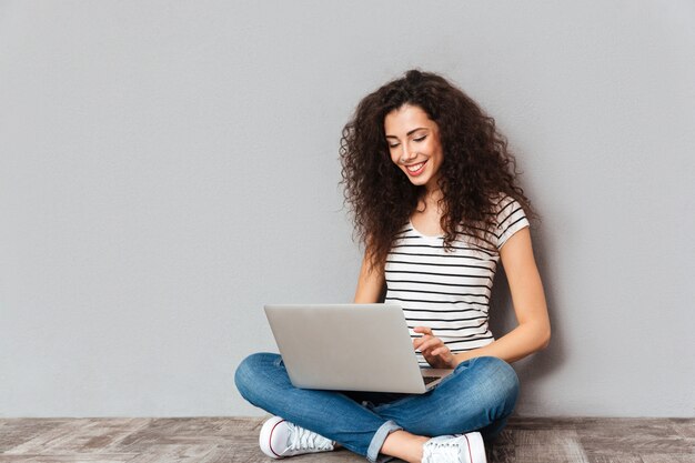 Het aantrekkelijke wijfje met mooie glimlach die in skype op elkaar inwerken of bericht typen die zilveren notitieboekje gebruiken terwijl het zitten in lotusbloem stelt op de vloer over grijze muur