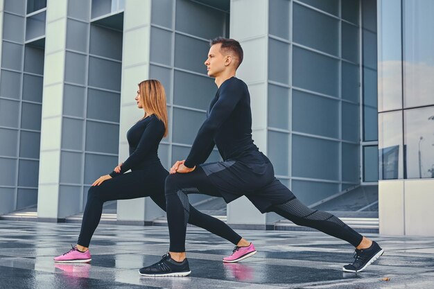 Het atletische fitnesspaar strekt zich uit over de achtergrond van het moderne gebouw. Volledig lichaamsbeeld.