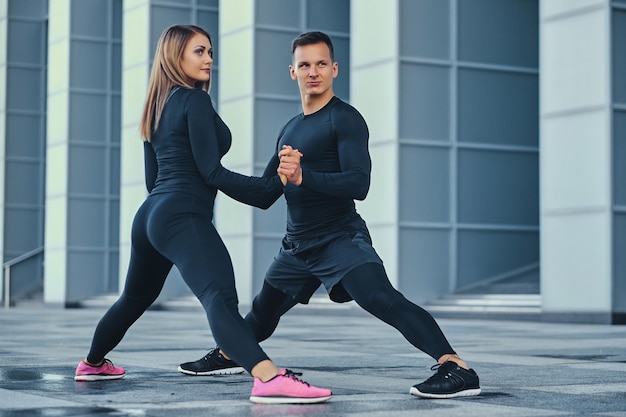 Het atletische fitnesspaar strekt zich uit over de achtergrond van het moderne gebouw. Volledig lichaamsbeeld.