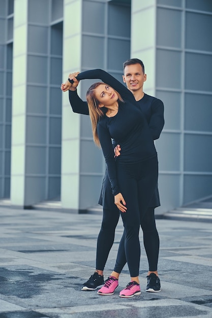 Het atletische fitnesspaar strekt zich uit over de achtergrond van het moderne gebouw. Volledig lichaamsbeeld.