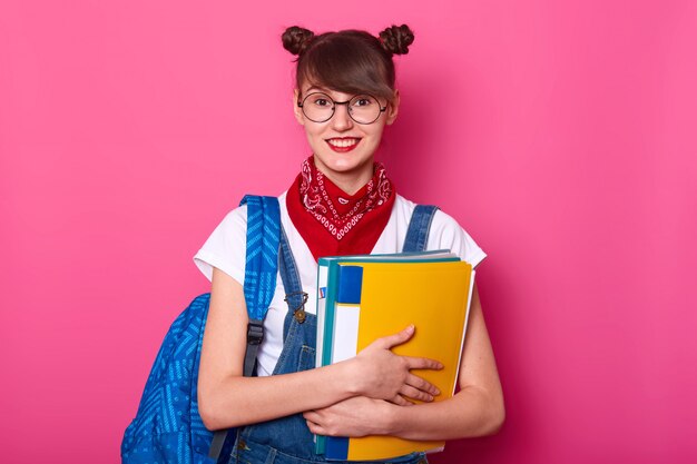 Het beeld van aanbiddelijk glimlachend schoolmeisje houdt kleurrijke bindmiddelen die op rooskleurige achtergrond worden geïsoleerd. Jong meisje met een gelukkige uitdrukking