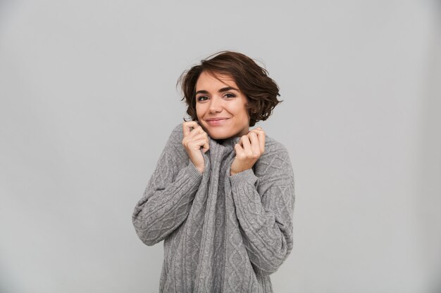 Het beeld van vrolijke jonge dame kleedde zich in sweater status geïsoleerd over grijze muur.