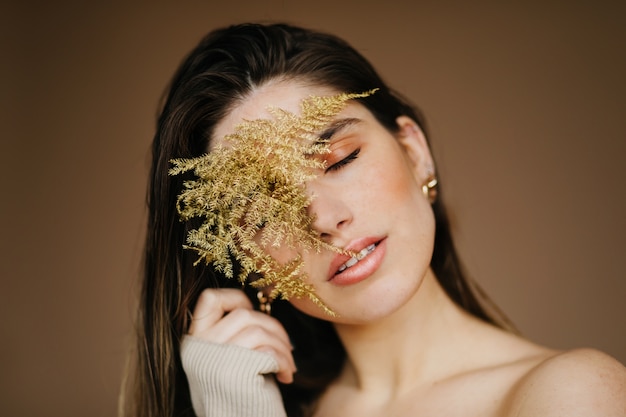 Het betoverende jonge vrouw stellen met gesloten ogen op bruine muur. goedgehumeurde, aangename dame met plant.