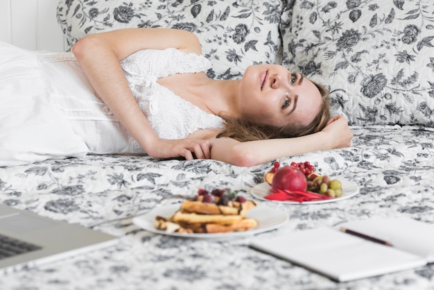 Het blonde jonge vrouw van het dagdromen die op bed met gezond ontbijt op bed liggen