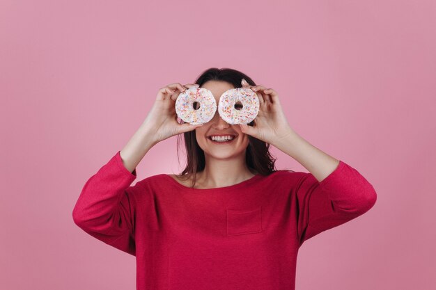 Het charmeren van jonge brunette houdt roze donuts vóór haar ogen die in de roze ruimte stellen