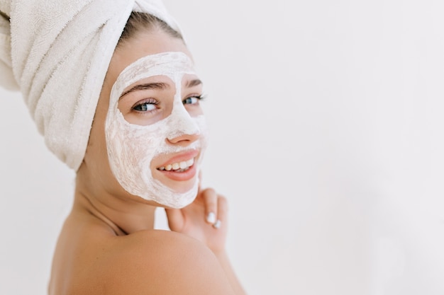 Het close-upportret van de mooie jonge vrouw die met handdoeken glimlacht na nemen bad maakt kosmetisch masker op haar gezicht.