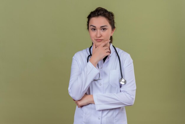 Het denkende arts jong meisje dat medische toga draagt die stethoscoop draagt, legde haar hand op wang op groene achtergrond
