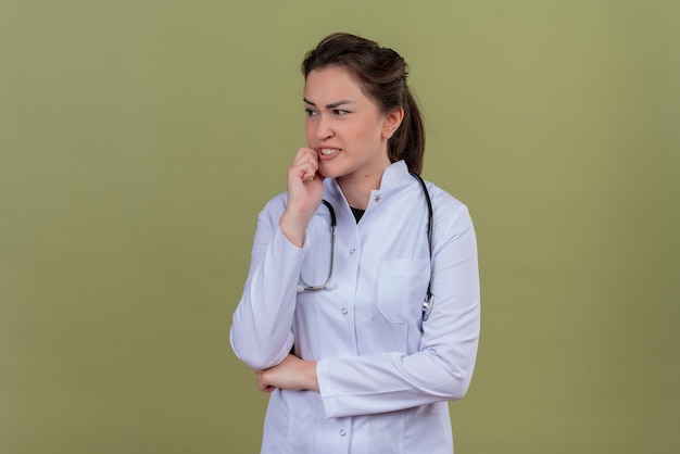 Het denkende arts jong meisje dat medische toga draagt die stethoscoop draagt, legde haar hand op wang op groene achtergrond
