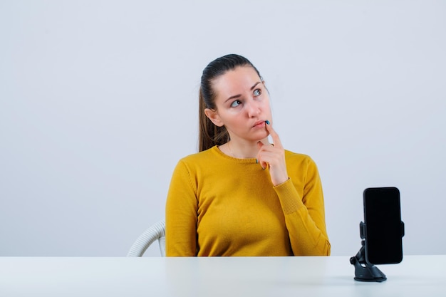 Het denkende bloggermeisje legt de wijsvinger op de lippen door voor de mobiele camera op een witte achtergrond te zitten