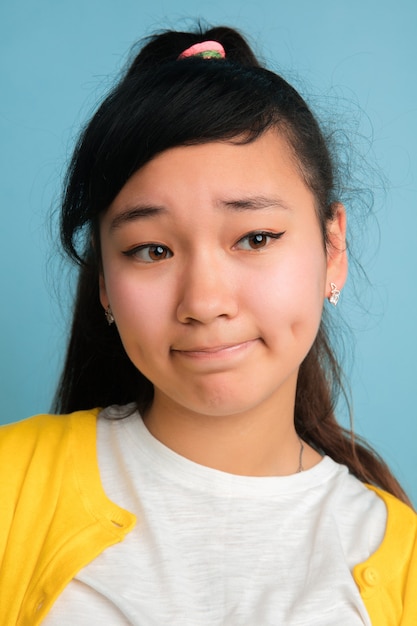 Het dichte omhooggaande portret van de Aziatische tiener dat op blauwe studioachtergrond wordt geïsoleerd. Mooi vrouwelijk donkerbruin model met lang haar. Concept van menselijke emoties, gezichtsuitdrukking, verkoop, advertentie. Ziet er verdrietig uit.