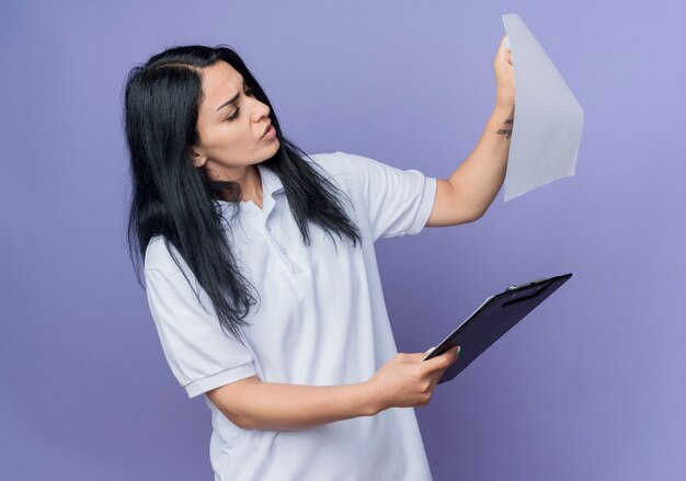 Het ernstige jonge donkerbruine Kaukasische meisje houdt klembord en bekijkt document blad dat op purpere muur wordt geïsoleerd