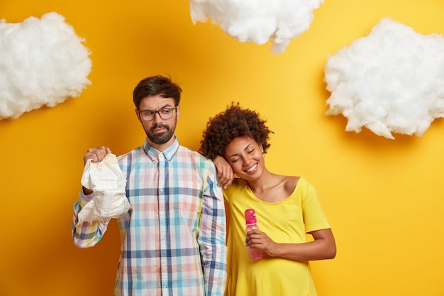 Gratis foto het familiepaar bereidt zich voor op de geboorte van een kind. zwangere vrouw en haar man poseren met babyfles en luier, klaar om binnenkort ouders te worden, kopen noodzakelijke dingen voor pasgeborenen, hebben leuke klusjes