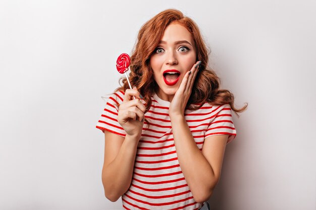 Het fascinerende gembermeisje stellen op witte muur met verbaasde glimlach. modieuze krullende vrouw met lolly.