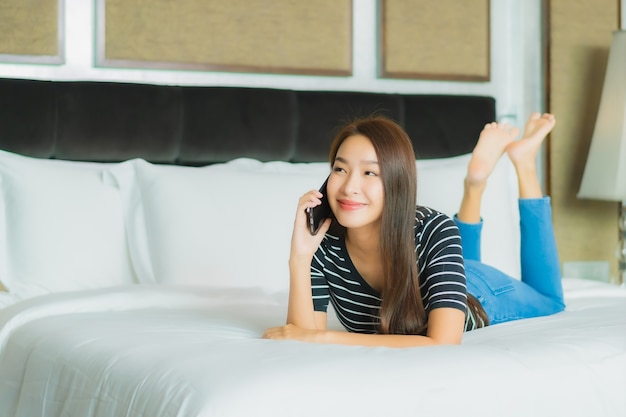 Het gebruik van slimme mobiele telefoon van de portret mooie jonge Aziatische vrouw op bed in slaapkamerbinnenland