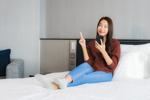 Het gebruik van slimme mobiele telefoon van de portret mooie jonge Aziatische vrouw op bed in slaapkamerbinnenland