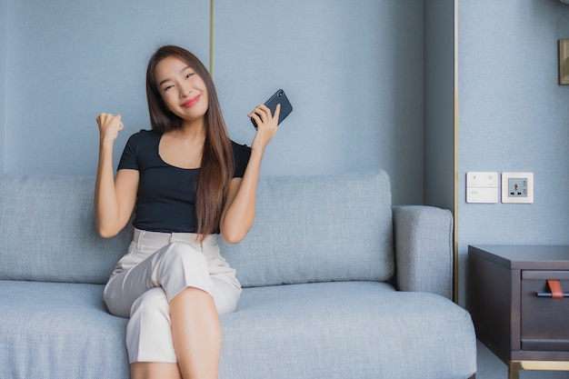 Het gebruiks slimme mobiele telefoon van de portret mooie jonge Aziatische vrouw op bank op woonkamergebied