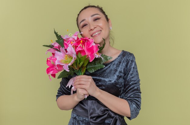 Het gelukkige Aziatische boeket van de vrouwenholding van bloemen glimlachen vrolijk kijkend naar voorzijde die de dag van internationale vrouwen viert die zich over groene muur bevindt