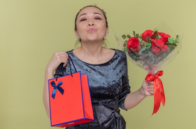 Het gelukkige Aziatische boeket van de vrouwenholding van rode rozen en papieren zak met cadeau blaast een kus die de dag van internationale vrouwen viert die zich over groene muur bevindt