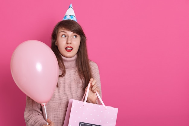 Het gelukkige donkerbruine meisje met roze ballon en bithday in handen aanwezig, houdt mond open, aangezien hij in verrassing is, kijkt opzij met verbaasd